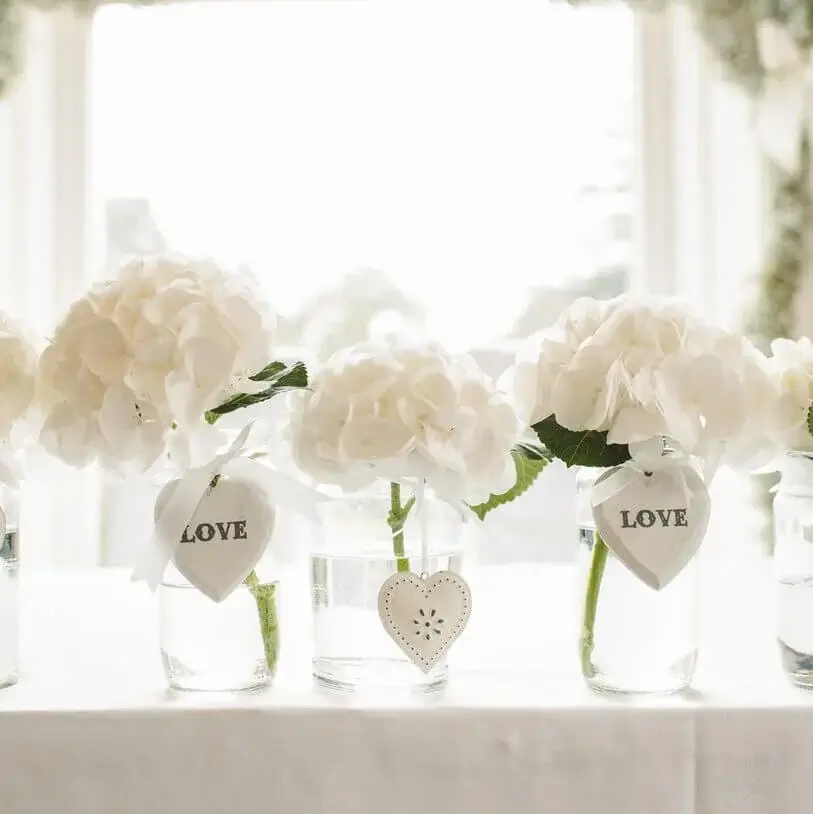 Photo of wedding table setting flower decorations, white flowers in jars filled with water with a white love heart decoration tied around the jars.